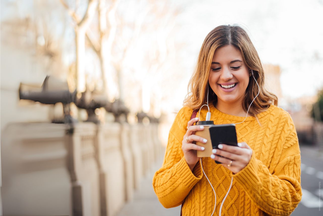 Junge Frau mit Kopfhörern sowie Kaffe und Smartphone in der Hand