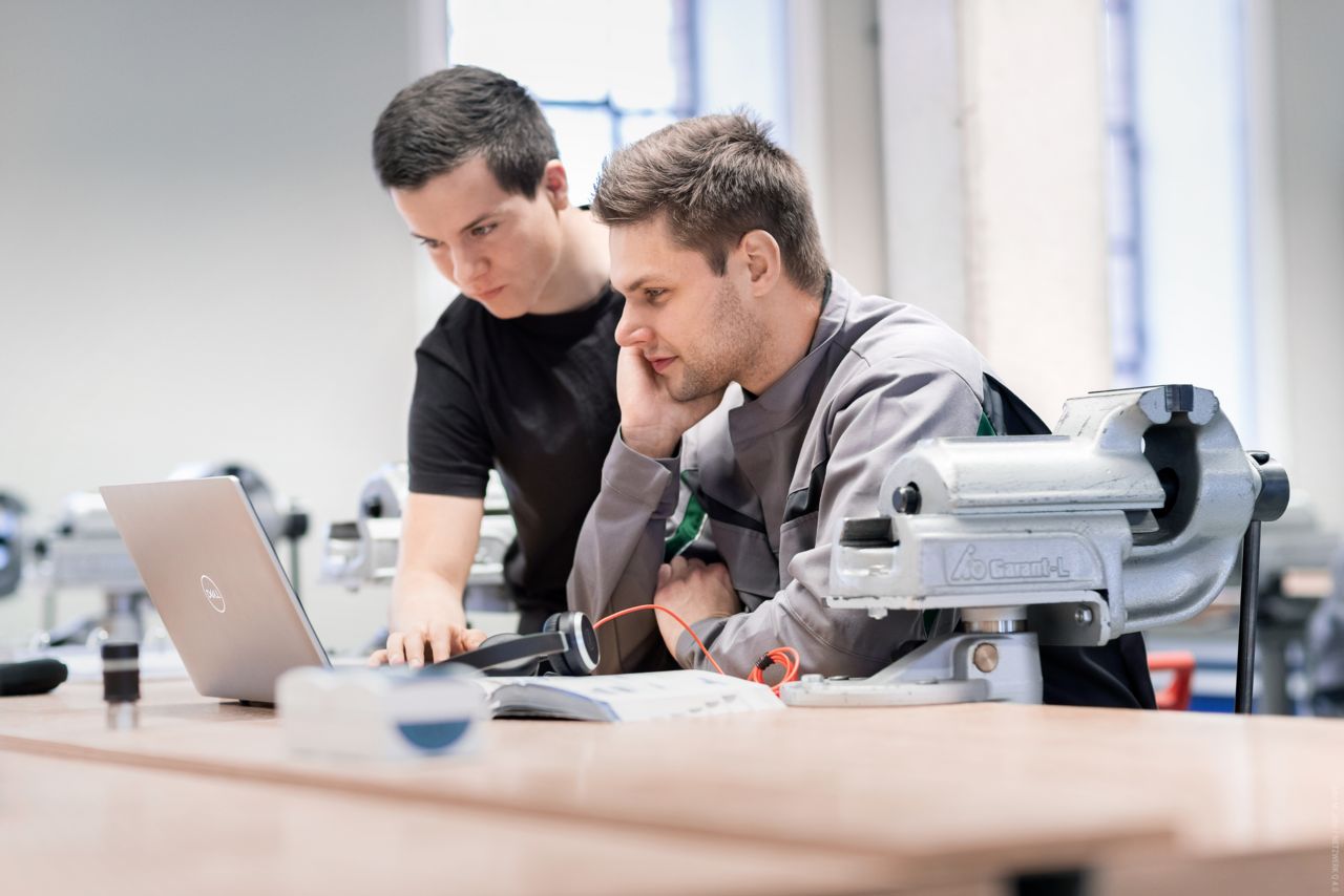 Azubi und Ausbilder schauen in einer Werkstatt gemeinsam auf einen Rechner